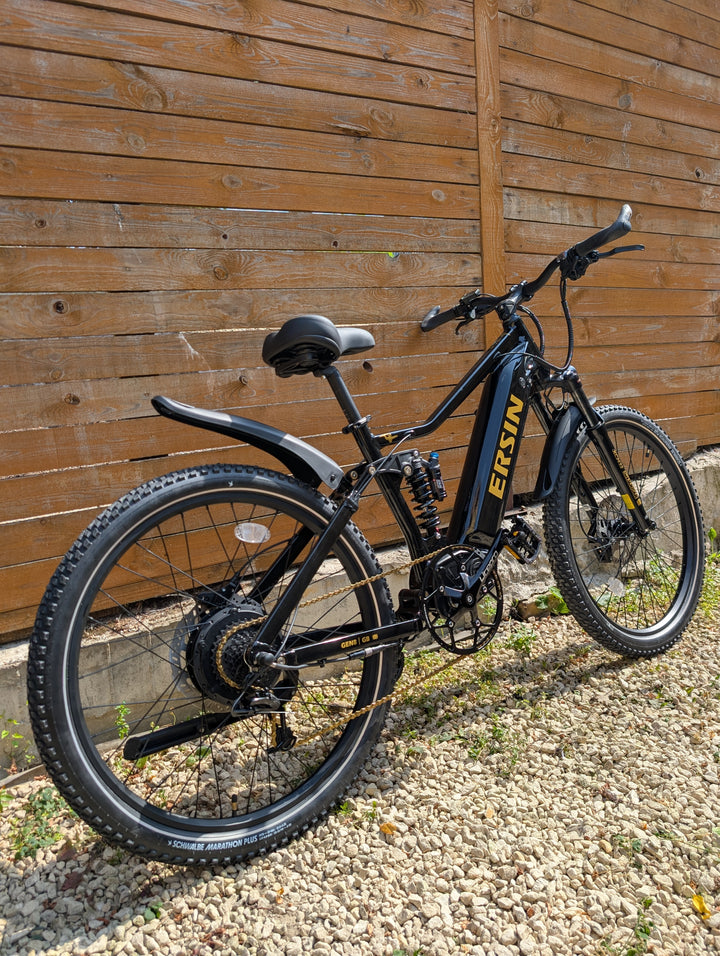 A black Ersin MTB electric mountain bike with gold branding, parked on gravel in front of a wooden fence.