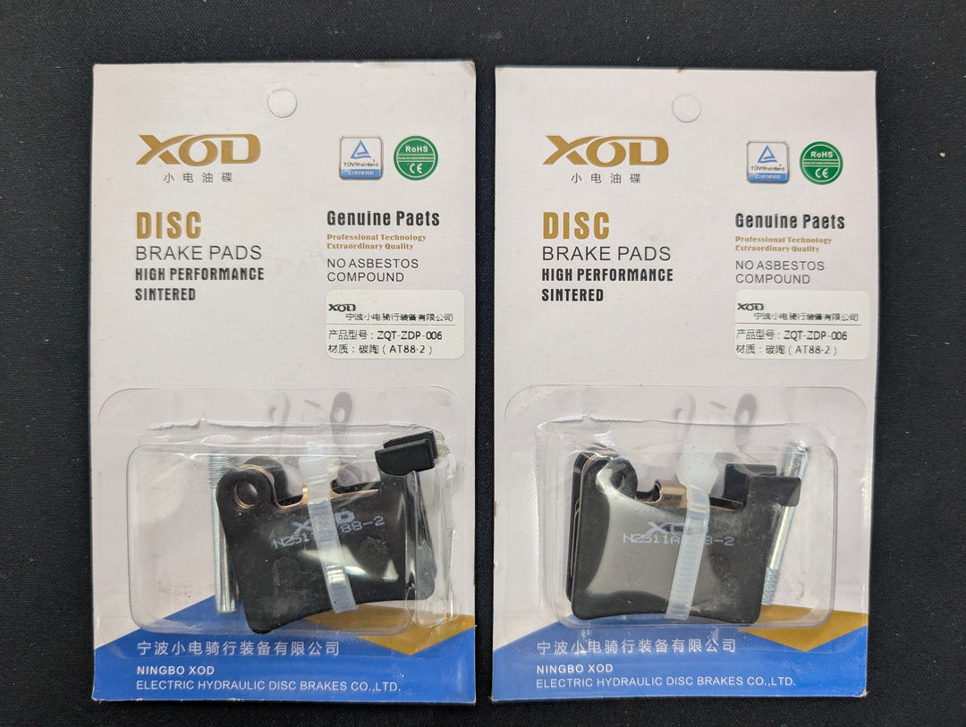 Two packaged XOD disc brake pads shown on a dark surface, each in a clear blister pack with printed retail card backing.