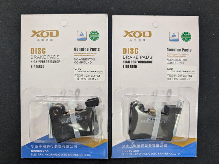 Two packaged XOD disc brake pads shown on a dark surface, each in a clear blister pack with printed retail card backing.